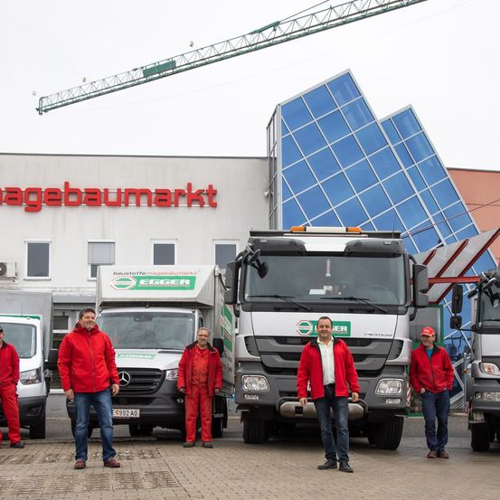 Eine Gruppe von Männern in roten Arbeitsanzügen steht vor Trucks vor einem Gebäude mit einem Kran darüber. Das Gebäude hat ein großes Schild mit der Aufschrift 'agebaumarkt'.
