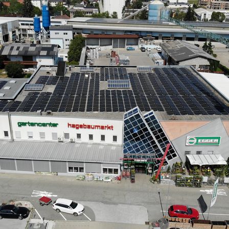 Eine Luftaufnahme eines Gartencenters mit Solardach und Parkplatz. Mehrere Autos sind geparkt, und ein roter Kran ist sichtbar.