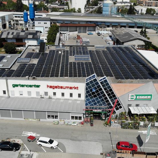 Eine Luftaufnahme eines Gartencenters mit Solardach und Parkplatz. Mehrere Autos sind geparkt, und ein roter Kran ist sichtbar.