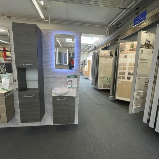 Eine moderne Badezimmerausstellung in einem Geschäft mit grauen Schränken, einem weißen Waschbecken und einem blauen Spiegel. Der Boden ist mit Teppich ausgelegt und die Wand ist aus Ziegeln mit einem blauen Schild.