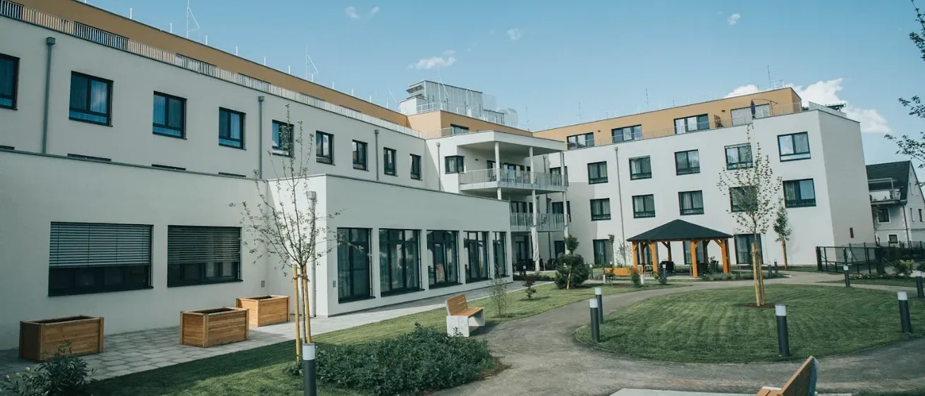 Ein modernes weißes Gebäude mit Glasfenstern und einem Weg, der zu einem Pavillon in einem Rasenbereich führt.