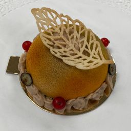 Ein wunderschön gestaltetes Dessert zeigt eine goldene Kugel, die mit einer blattartigen Dekoration, roten Beeren und kleinen Früchten verziert ist und auf einem silbernen Teller serviert wird.