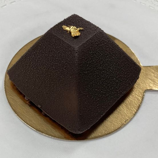 Ein pyramidenförmiger Schokoladenkuchen mit einer goldenen Biene darauf, der auf einem goldenen Teller auf einer weißen Oberfläche ruht.