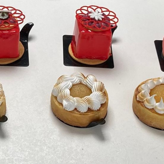 Fünf Mini-Desserts sind auf einer weißen Oberfläche ausgestellt. Zwei sind rot mit dekorativen Toppings und drei sind mit Sahne gekrönt. Jedes Dessert steht auf einem kleinen schwarzen Ständer.