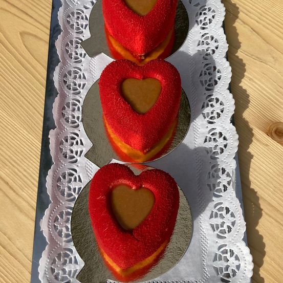 Vier herzförmige Kuchen mit roter Glasur und Karamellfüllung auf einem silbernen Teller mit Spitzenbordüre auf einer Holzoberfläche.