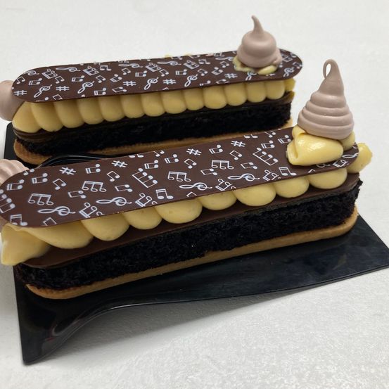 Zwei Schokoladenüberzogene Desserts mit musikalischen Noten-Designs stehen auf einer schwarzen Oberfläche. Jedes Dessert hat eine Schicht gelber Creme und schokoladebedeckte Cremepuffs.