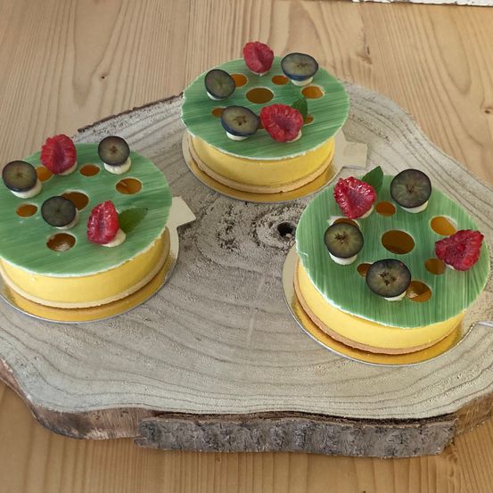 Drei Mini-Kuchen stehen auf einem Holzbrett, jeder mit gelber Creme und grüner Glasur. Sie haben dekorative Früchte wie Himbeeren und Pflaumen.