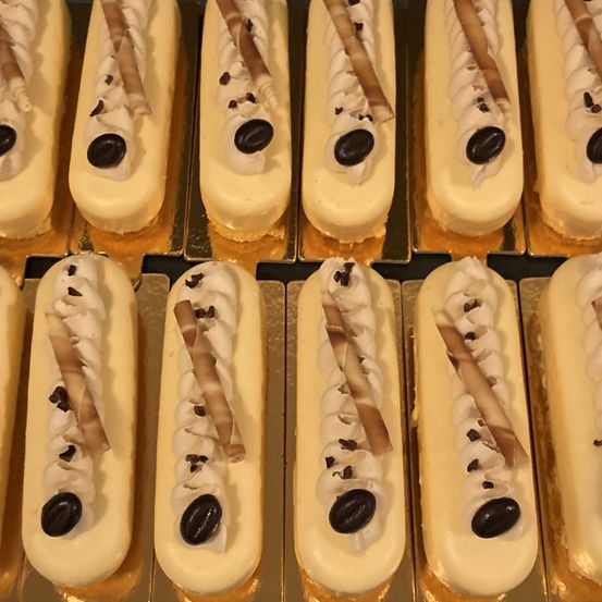 Mehrere längliche Gebäckstücke mit einer cremigen Füllung, überzogen mit weißer Creme und braunen Stäbchen, dekoriert mit Kaffeebohnen.