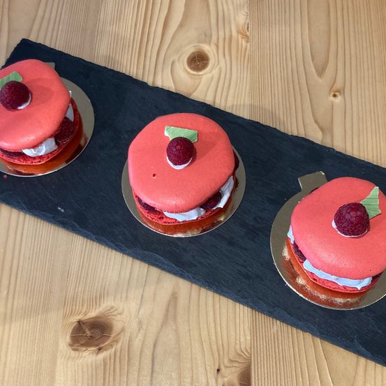 Drei rote Samtkuchen mit weißer Creme und Himbeeren auf einem schwarzen Schiefertablett, auf einer Holzoberfläche platziert.