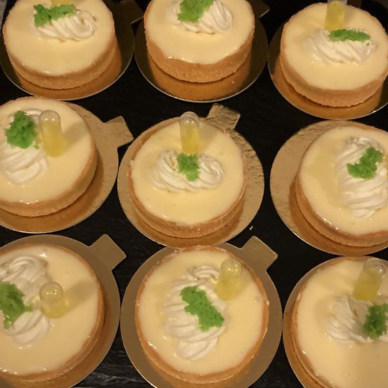 Mehrere kleine Törtchen mit weißer Sahne und grünen Blättern darauf sind auf einer schwarzen Oberfläche angeordnet. Sie sind mit einer kleinen Flasche gelber Flüssigkeit gekrönt.