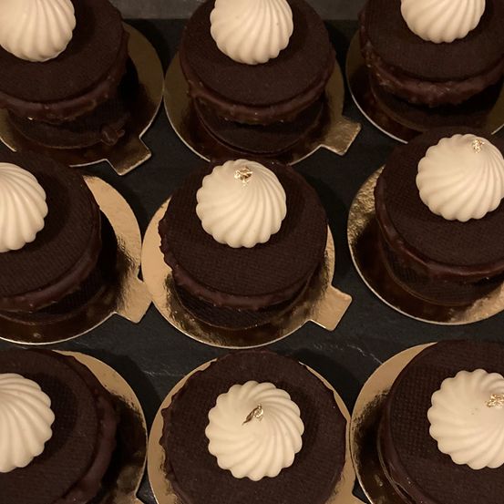 Eine Nahaufnahme von mehreren Schokoladenkuchen mit weißer Creme obenauf, jeweils verziert mit einem goldenen Blatt auf der Creme, auf goldumrandeten Tellern auf einer dunklen Oberfläche.