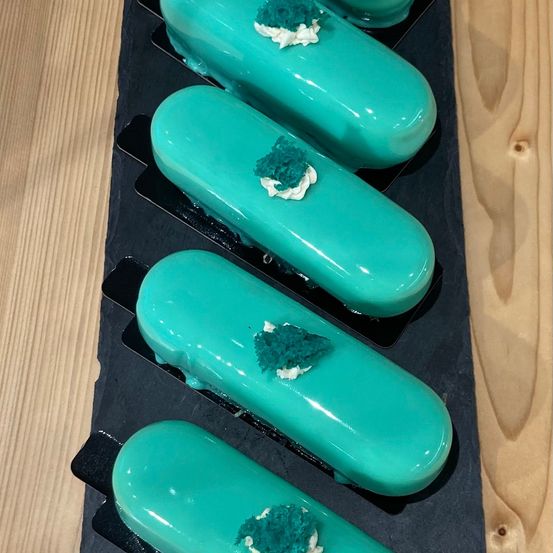 Fünf türkisfarbene Dessertartikel sind auf einem schwarzen Tablett angeordnet, das auf einer Holzoberfläche platziert ist. Jedes hat weiße Creme und blaue dekorative Elemente.