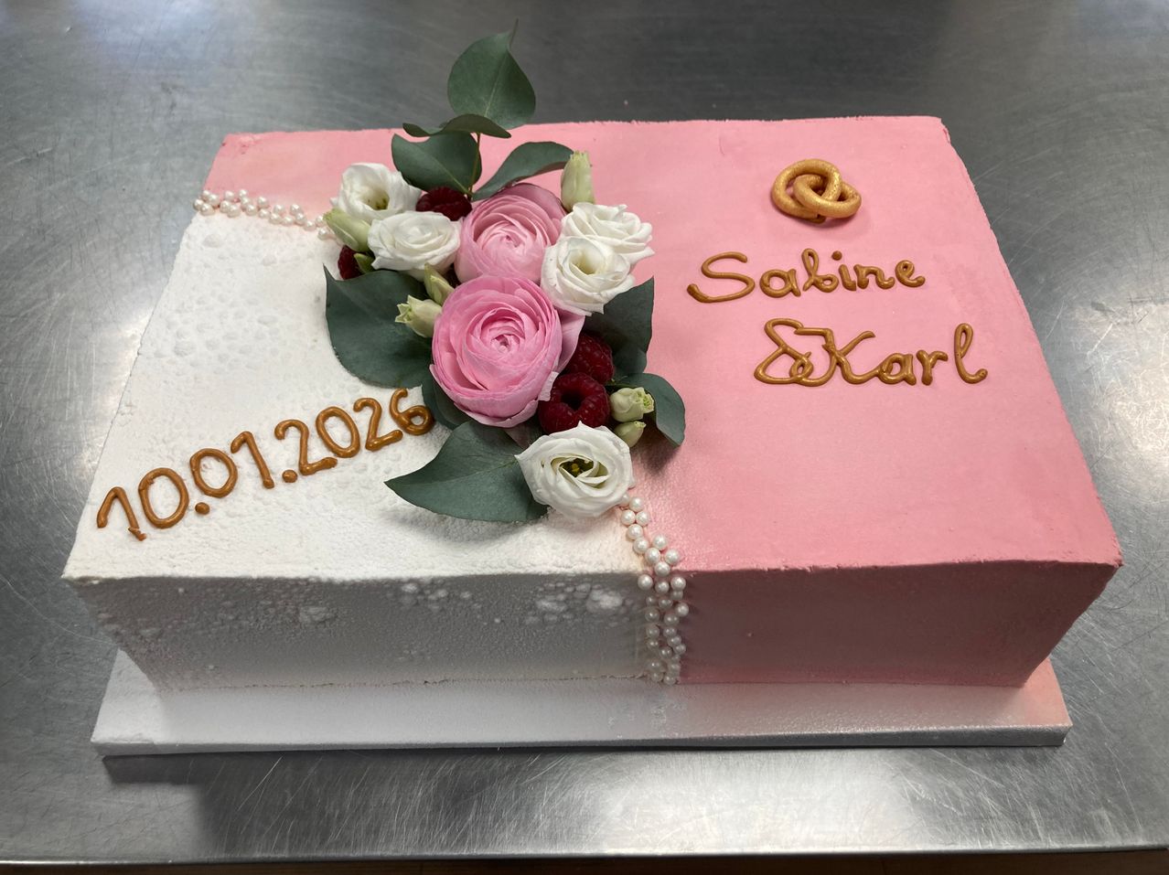 Ein rosa und weißer Kuchen mit Blumen, Perlen und goldenen Brezeln trägt die Aufschrift '01.07.2026' und 'Sabine & Karl'. Der Kuchen steht auf einer weißen Oberfläche.