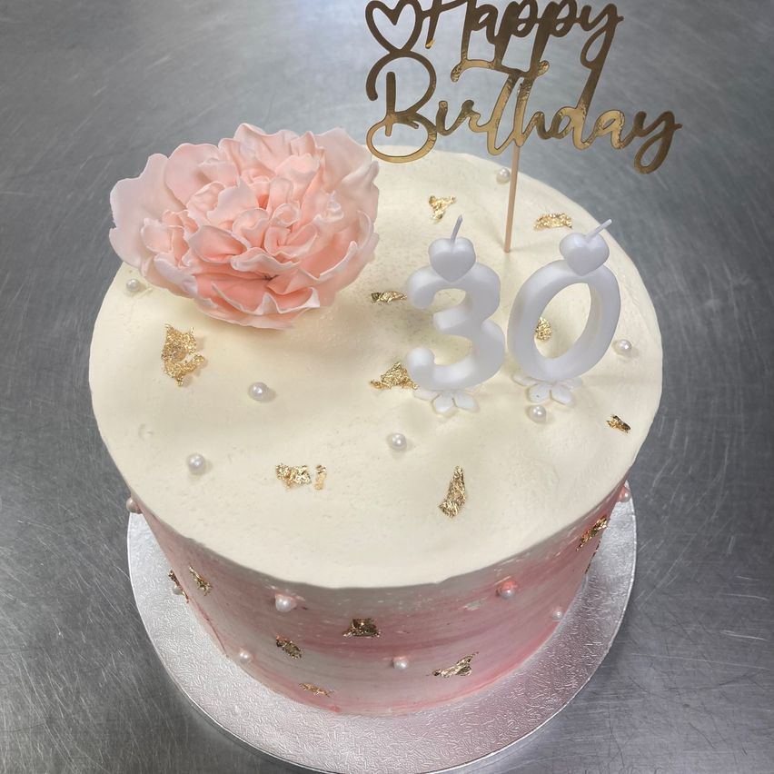 Ein Geburtstagskuchen mit weißer Glasur, rosa Akzenten und goldenen Dekorationen. Er hat eine rosa Blume und einen goldenen 'Happy Birthday'-Schriftzug oben drauf. Der Kuchen hat zwei Kerzen, '30' auf der Oberseite.
