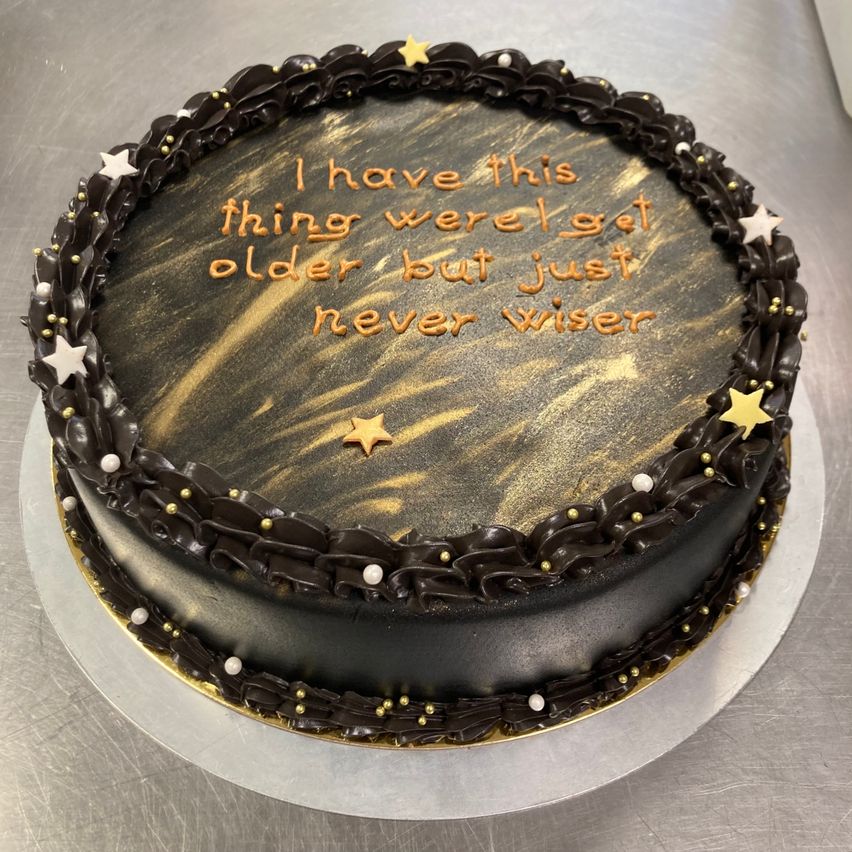 Ein Schokoladenkuchen mit schwarzem und goldenem Muster und einer dekorativen Einfassung, verziert mit Sternen. Der Kuchen trägt den Schriftzug 'Ich habe dieses Ding, bei dem wir älter werden, aber einfach nie klüger.'