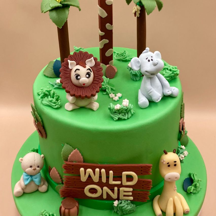 Ein leuchtend grüner Kuchen mit Dschungeltieren wie Löwe, Elefant, Bär und Giraffe. Ein Holzschild sagt 'WILD ONE'. Grüne Pflanzen und Blumen umgeben den Kuchen.