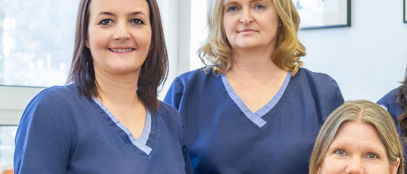 Vier Frauen in blauen Kitteln stehen in einer Zahnarztpraxis und lächeln für die Kamera. Sie tragen Halsketten und Ohrringe. Ein roter Stuhl und ein gerahmtes Bild schmücken den Hintergrund.