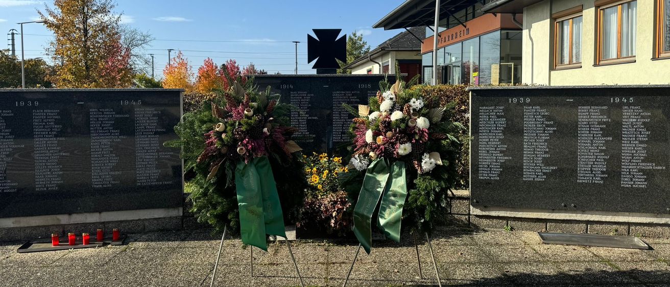 Zwei Kränze mit Bändern stehen vor Gedenktafeln, umgeben von Grünpflanzen und Blumen. Die Tafeln sind mit Daten und Namen graviert, möglicherweise zur Erinnerung an gefallene Soldaten. Ein Gebäude mit braunem Dach und Glasfenstern ist im Hintergrund zu sehen.