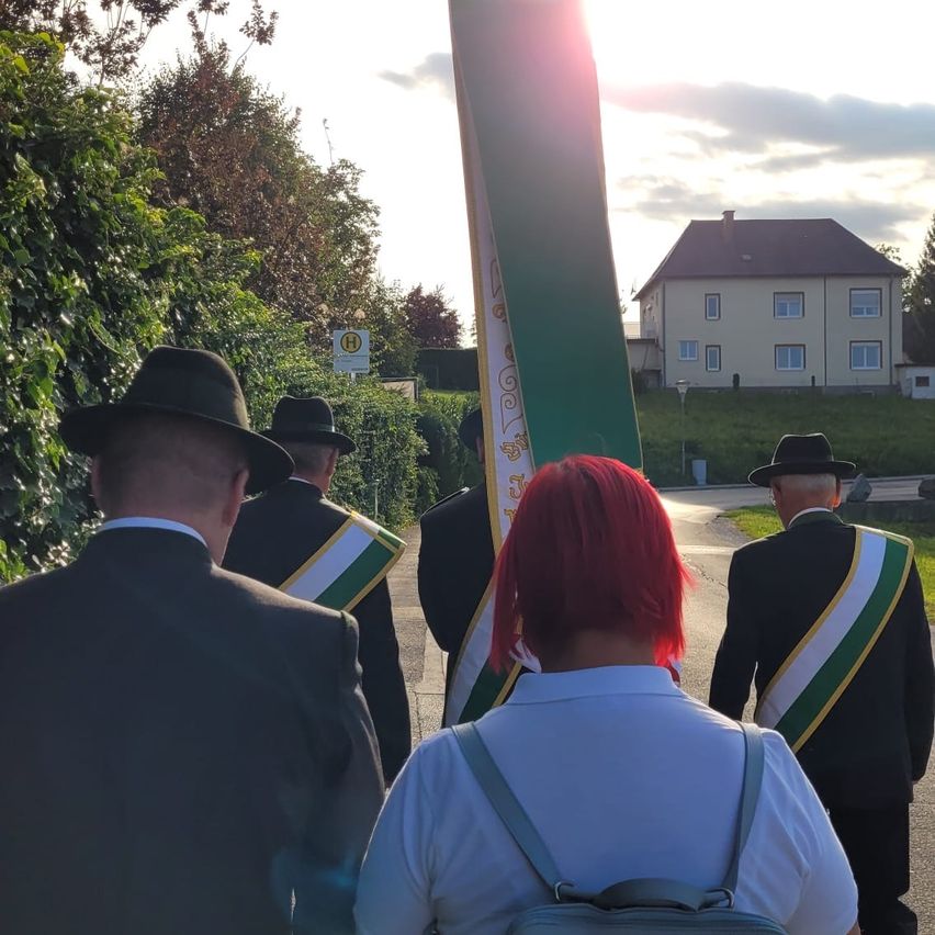 Eine Gruppe formell gekleideter Personen geht zusammen auf einer Straße, mit einem Haus und Grünpflanzen im Hintergrund.