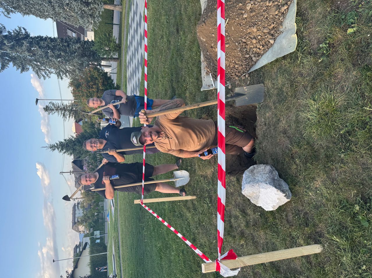 Vier Männer benutzen Schaufeln, um ein Loch in einem Grasbereich mit einer rot-weiß gestreiften Barriere zu graben. Ein Mann trägt ein braunes Shirt, und ein anderer trägt ein schwarzes Shirt. In der Nähe befindet sich ein großer Stein.