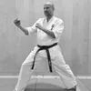 Ein Mann in einem weißen Karate-Anzug mit einem schwarzen Gürtel führt eine Kampfkunstpose durch, steht auf einem Mattenboden mit gespreizten Beinen und seinen Händen vor sich positioniert.