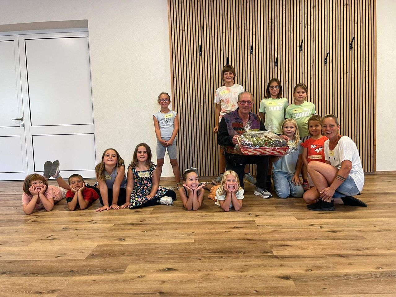 Eine Gruppe von Kindern und Erwachsenen posiert für ein Foto in einem Raum. Ein Mann mit einem Geschenkkorb sitzt in der Mitte, einige Kinder sitzen und andere stehen hinter ihm. Der Raum hat Holzwände.