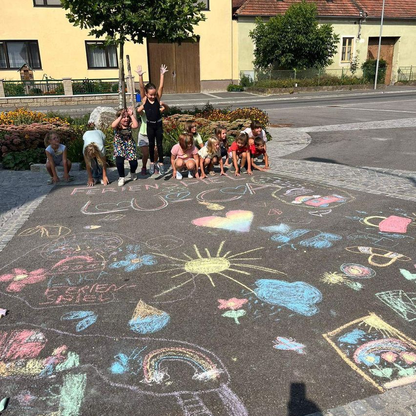 Eine Gruppe von Kindern malt farbenfrohe Kreidekunst auf der Straße, darunter eine Sonne, Blumen und Regenbögen. Sie hocken und stehen hinter der Kunst, mit Gebäuden und Grünpflanzen im Hintergrund.