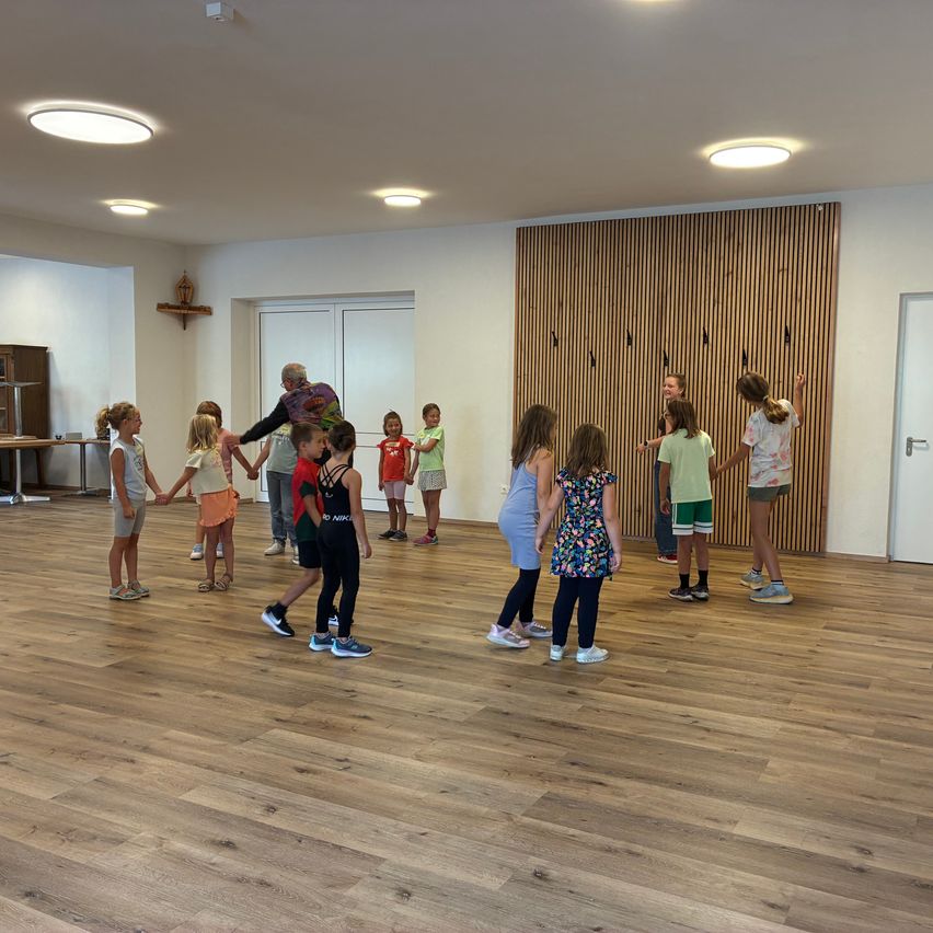 In einem gut beleuchteten Raum mit Holzboden führen mehrere Kinder und ein Erwachsener eine Aktivität durch. Die Kinder tragen lässige Sommerkleidung und Turnschuhe. Der Erwachsene, in einem bunten Shirt gekleidet, scheint sie zu leiten. Der Raum verfügt über eine Holzwand, Deckenleuchten und eine Tür im Hintergrund.