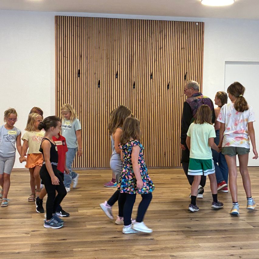 Eine Gruppe von Kindern und zwei Erwachsenen versammelt sich in einem Raum mit Holzboden. Sie halten Händchen und praktizieren möglicherweise eine Tanzroutine. Hinter ihnen befindet sich eine Holzwand mit Metallhaken.