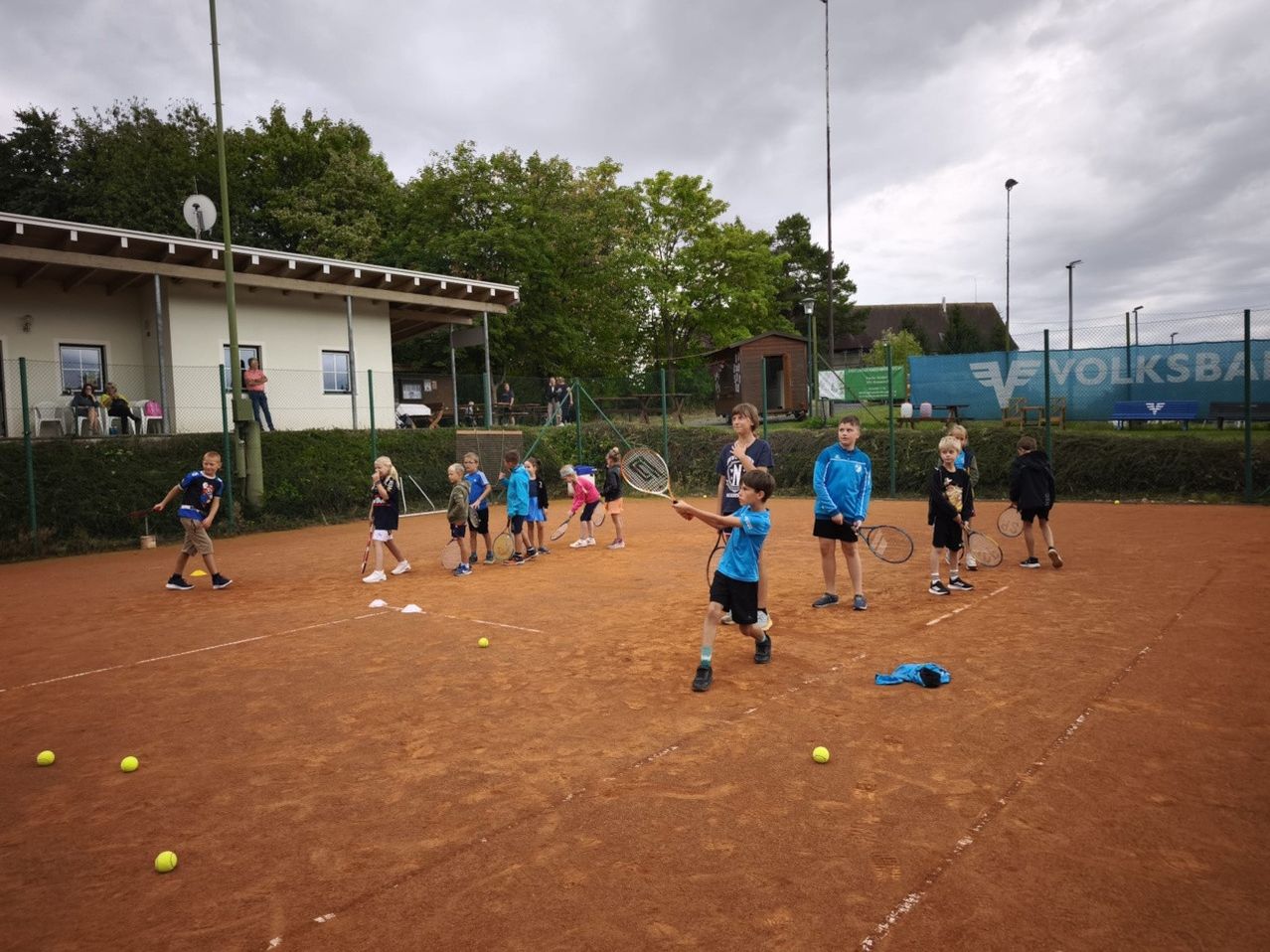Eine Gruppe von Kindern übt Tennis auf einem Außenplatz bei bewölktem Himmel. Ein Junge schlägt einen Tennisball.