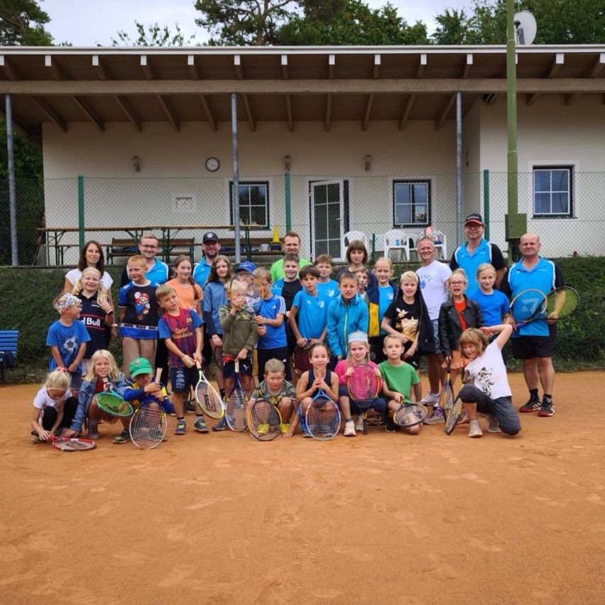 Eine Gruppe von Kindern und Erwachsenen mit Tennisschlägern posiert auf einem Sandplatz mit einem Gebäude im Hintergrund.