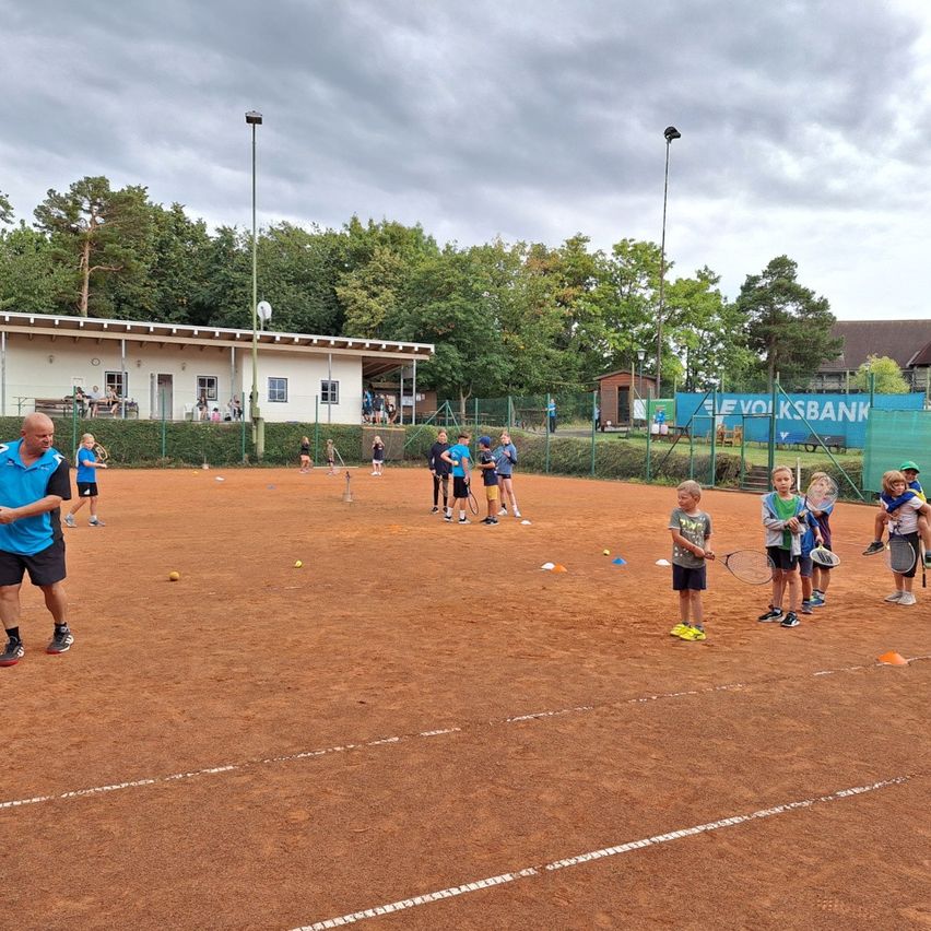 An einem bewölkten Tag üben Kinder Tennis auf einem Sandplatz. Orangefarbene Kegel sind um den Platz herum aufgestellt.
