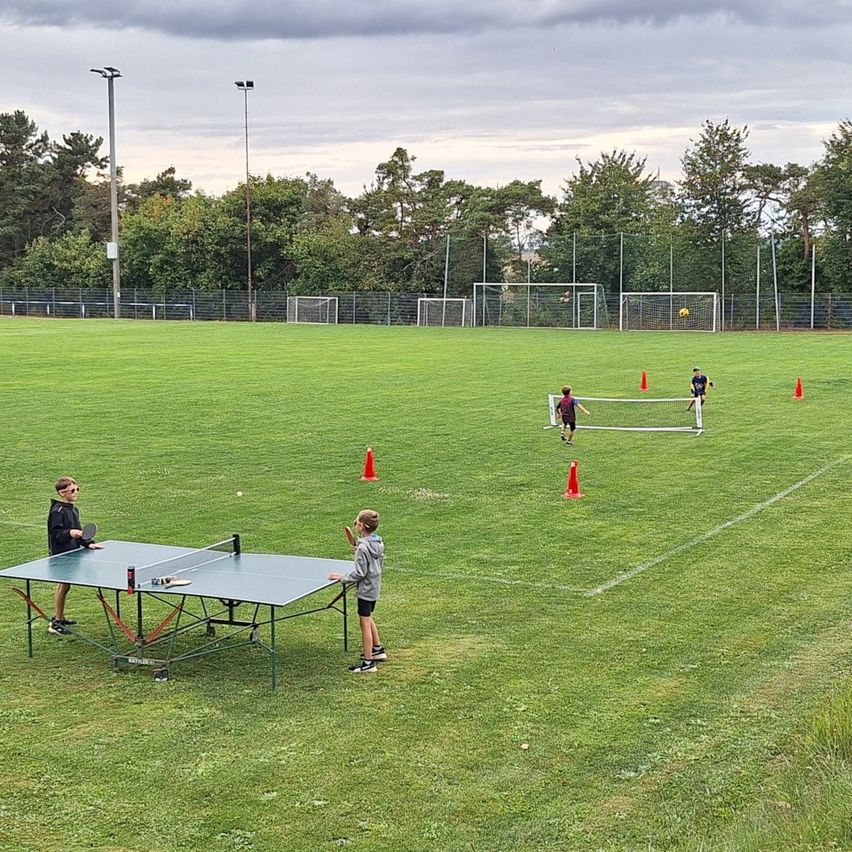 Zwei Kinder spielen Tischtennis auf einem Rasenfeld, umgeben von orangefarbenen Hütchen. Im Hintergrund befinden sich Fußballtore und Bäume.