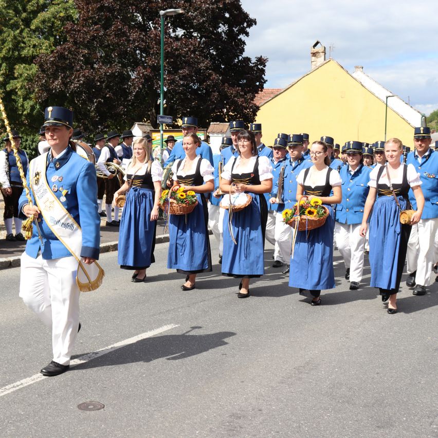 Eine Musikkapelle zieht mit Instrumenten bewaffnet die Straße entlang und trägt traditionelle Kostüme. Einige halten Blumenkörbe.