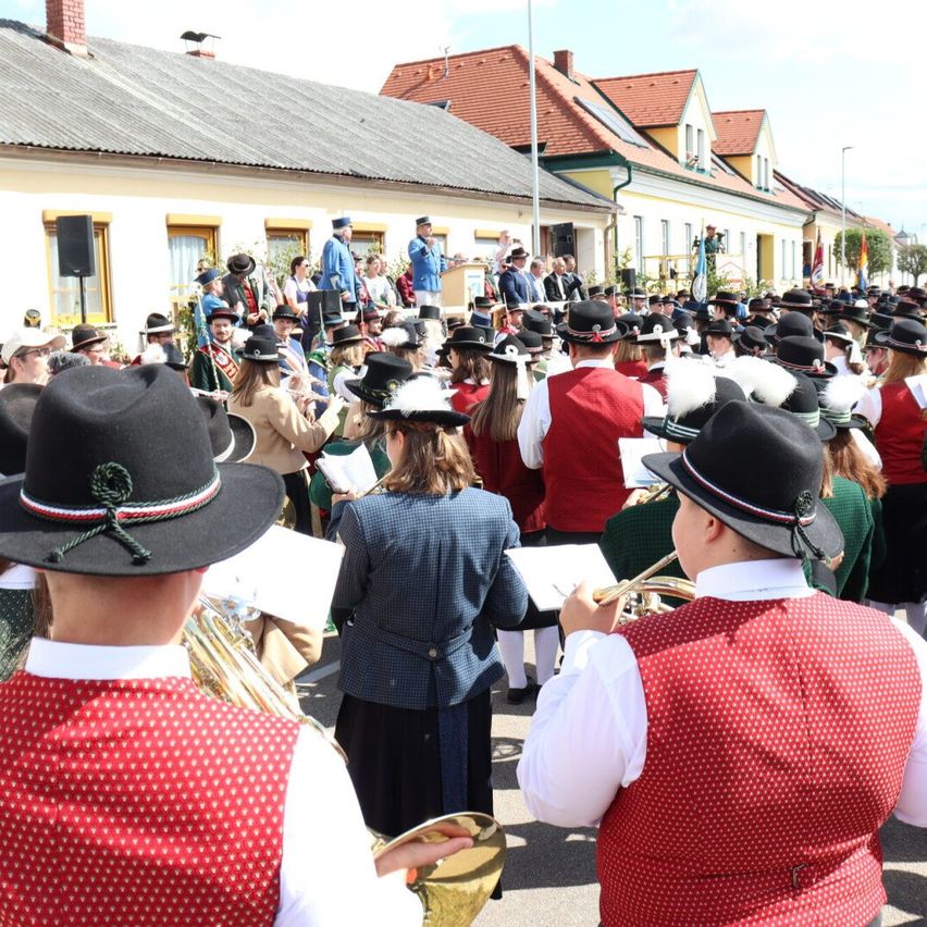Eine Blaskapelle spielt vor einer Menge in traditioneller Kleidung. Einige Musiker spielen Blechblasinstrumente, während andere Notenblätter halten. Das Publikum ist vor einem Gebäude mit roten Dächern versammelt.