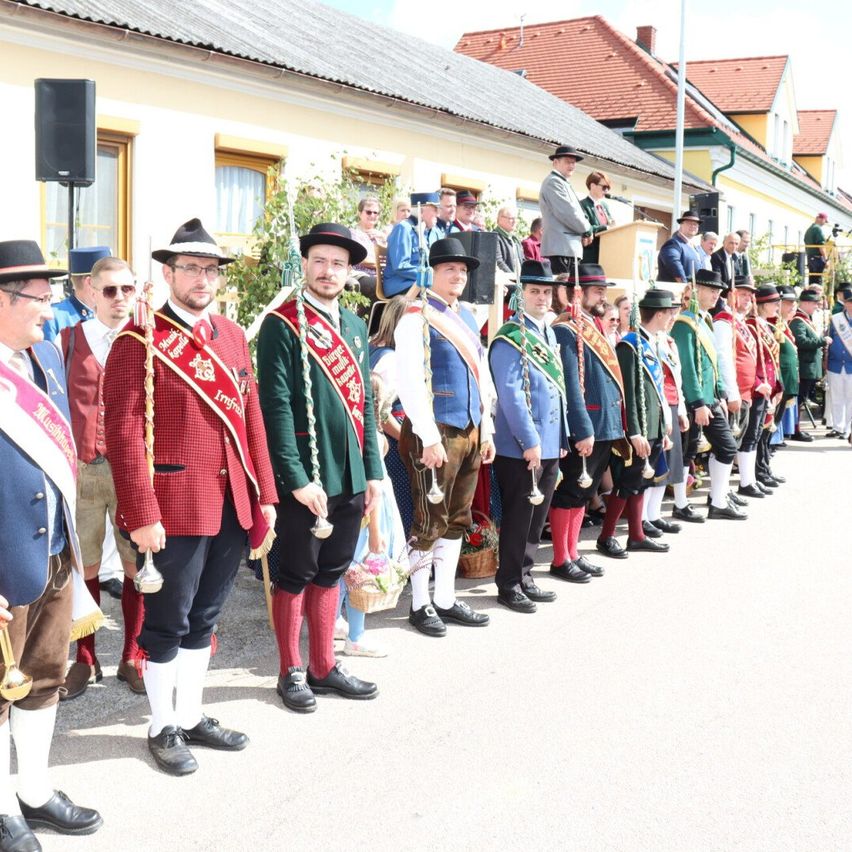 Eine Gruppe von Männern in traditionellen Uniformen steht in einer Reihe, hält Glocken und Blumen. Ein Gebäude mit rotem Dach steht hinter ihnen, und ein Lautsprecher befindet sich auf der linken Seite.