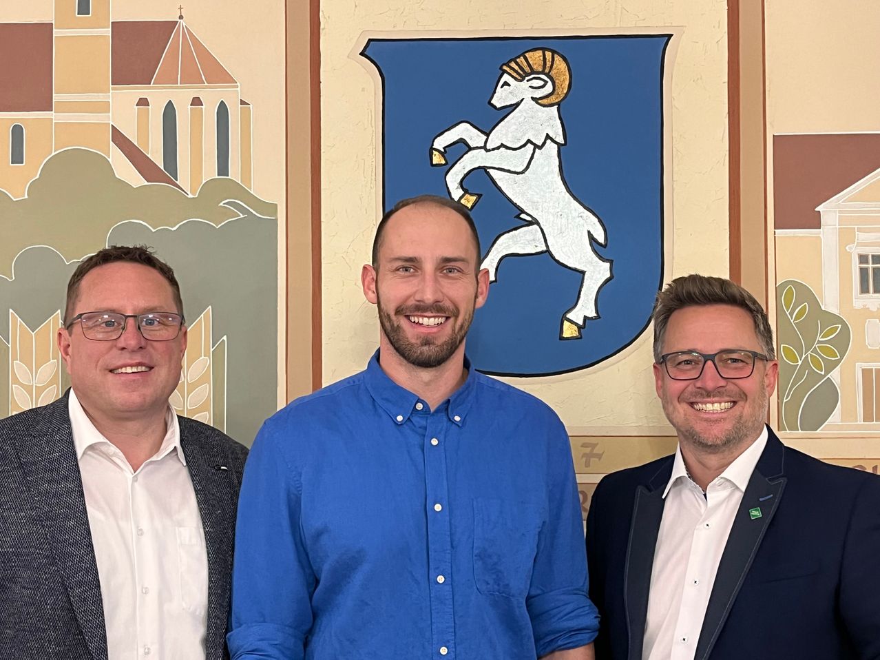 Drei Männer stehen gemeinsam vor einer Wandmalerei mit einer Kirche und einem Wappen mit einem Widder.