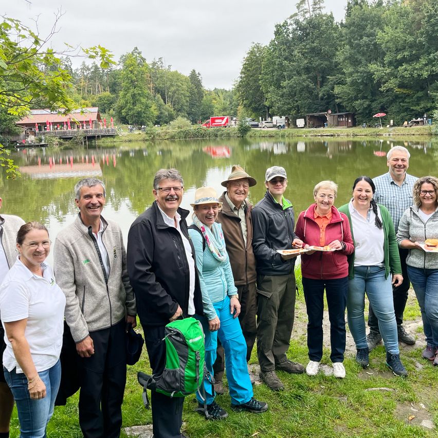 Eine Gruppe von Menschen lächelt und posiert für ein Foto in der Nähe eines Sees. Sie tragen verschiedene Kleidungsstücke und einige halten Teller mit Essen. Hinter ihnen steht ein Gebäude mit einem roten Dach und einer Brücke. Bäume und eine Rasenfläche sind ebenfalls vorhanden.