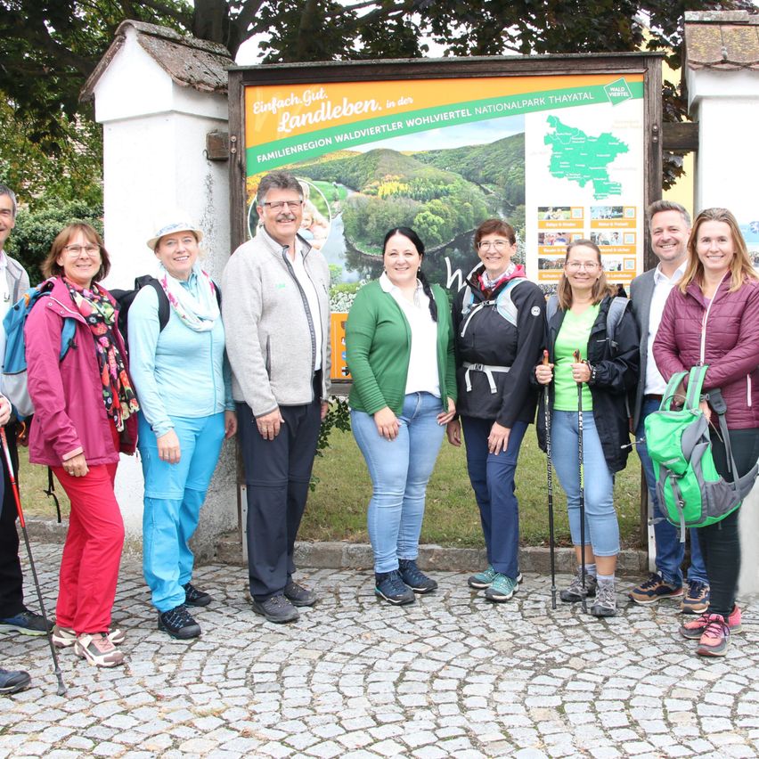 Eine Gruppe von Menschen steht vor einem Schild für einen Wanderweg. Sie tragen verschiedene Outdoor-Kleidung und haben Rucksäcke dabei. Das Schild zeigt eine Karte und Text in einer fremden Sprache.