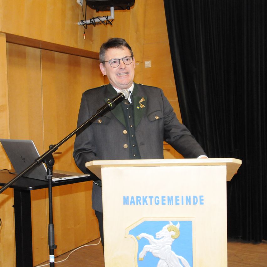 Ein Mann in einem formellen Anzug steht an einem Podium mit einem Mikrofon. Hinter ihm befindet sich ein Laptop auf einem Tisch, und ein Vorhang ist teilweise zugezogen. Das Podium trägt den Text 'MARKTGEMEINDE' und ein Symbol eines Widders.