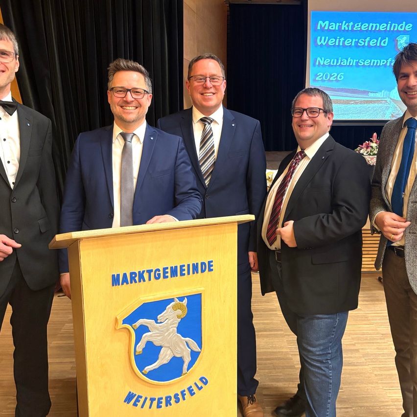 Fünf Männer in Anzügen stehen lächelnd für ein Foto an einem Podium mit einem Logo. Dahinter zeigt eine Projektionsleinwand eine Präsentation mit dem Titel 'Marktgemeinde Weitersfeld Neujahrsempfa 2026.'