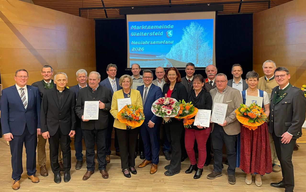 Eine Gruppe von Menschen in formeller Kleidung posiert mit Blumen und Zertifikaten für ein Foto, während eine Projektionsleinwand im Hintergrund 'Marktgemeinde Weitersdorf Neujahrsempfang 2026' anzeigt.