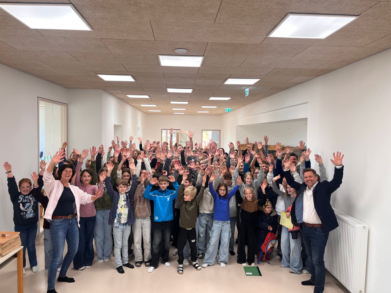 Eine Gruppe von Menschen, darunter Kinder, posiert für ein Foto in einem Raum mit fluoreszierenden Deckenleuchten. Die Erwachsenen lächeln und heben ihre Hände.