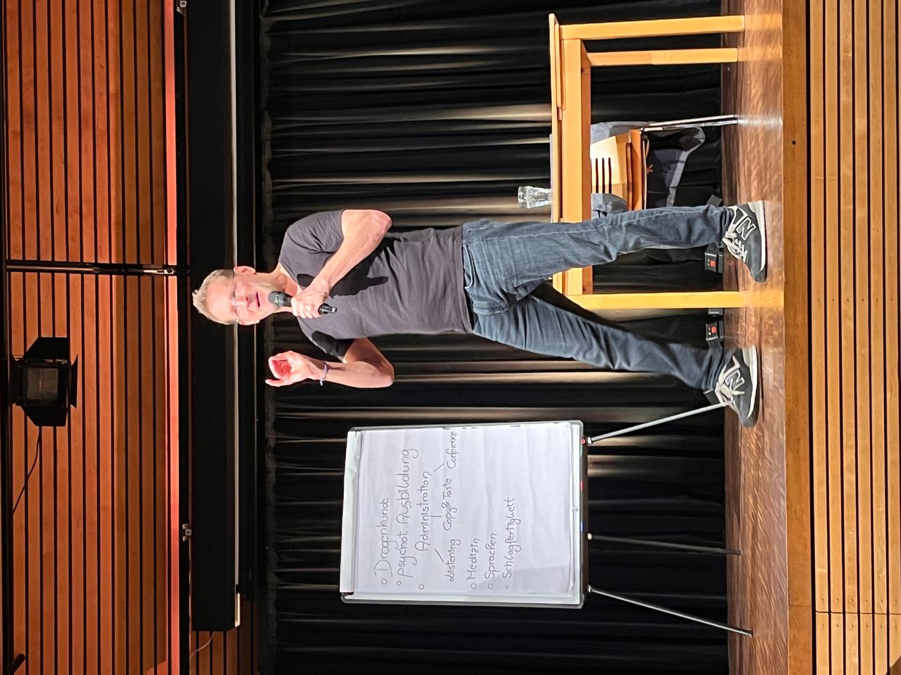 Ein Mann steht auf der Bühne, hält ein Mikrofon in der Hand und streckt sich über eine Tafel mit einer Liste von Namen und Themen. Die Bühne hat Holzwände und Vorhänge im Hintergrund.