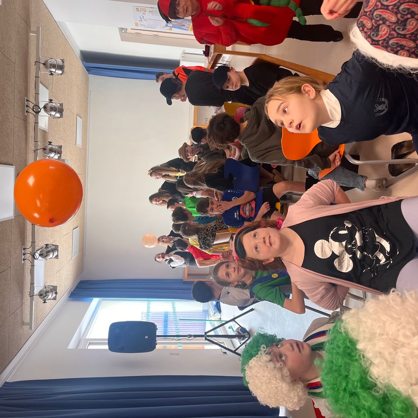 Viele Kinder in Kostümen versammeln sich in einem Raum. Ein großer orangefarbener Ballon ist vorhanden. Es gibt blaue Vorhänge und an der Wand montierte Lampen.