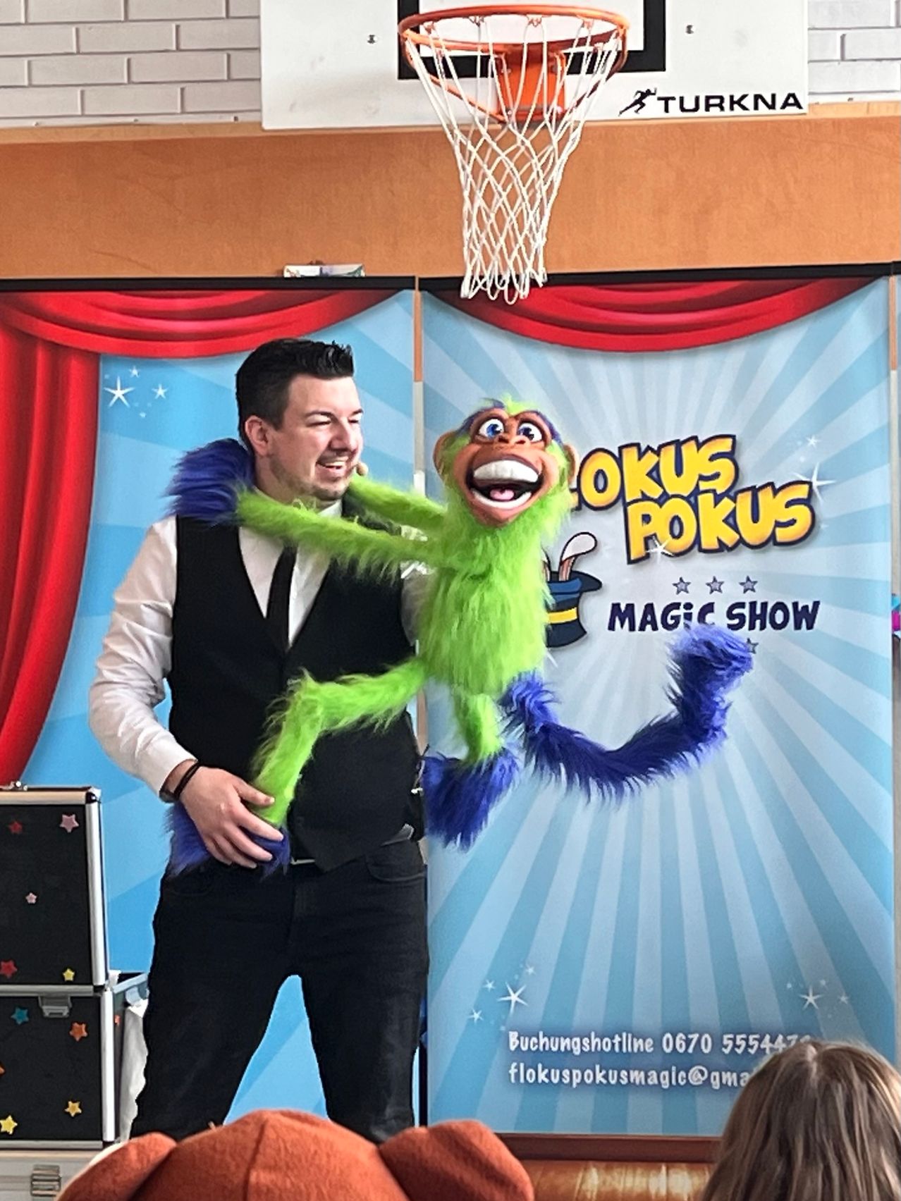 Ein Mann in Anzug und Krawatte hält eine grüne Puppe mit blauem Schwanz und Fell. Die Puppe lächelt und scheint eine Zaubershow aufzuführen.