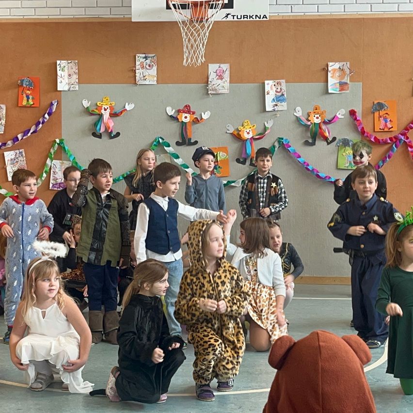Eine Gruppe von Kindern in verschiedenen Kostümen tritt in einer Turnhalle mit einem Basketballkorb und Wanddekorationen auf.