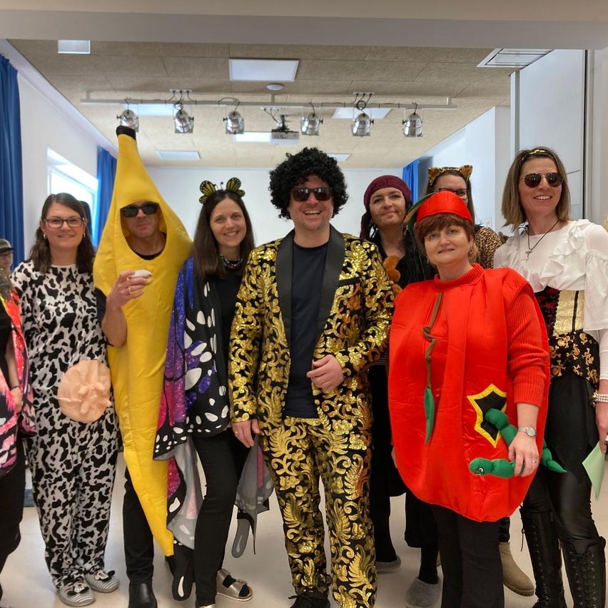 Eine Gruppe von Menschen in Kostümen posiert für ein Foto in einem Flur. Eine Person trägt eine Bananen-Verkleidung, eine andere eine Tomaten-Verkleidung und andere tragen verschiedene Kostüme.