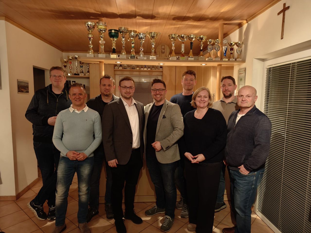 Eine Gruppe von Menschen, bestehend aus Männern und Frauen, posiert für ein Foto. Sie stehen nebeneinander, und einige von ihnen lächeln. Hinter ihnen sind Trophäen und Auszeichnungen auf einem Regal ausgestellt.