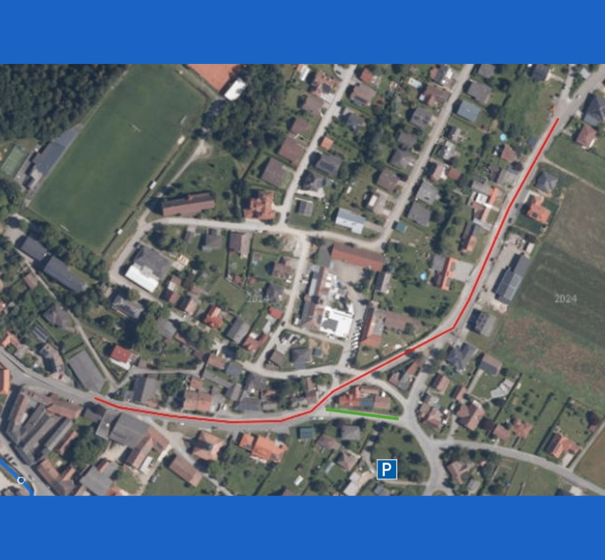 Luftaufnahme einer kleinen Stadt mit einer roten Linie, die einen Weg oder eine Straße anzeigt. Gebäude und Häuser sind um ein zentrales grünes Feld herum verstreut.